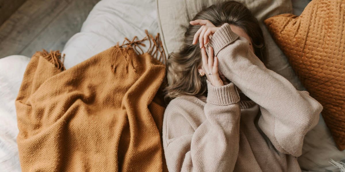 A white woman with brown hair and her face obscured by her hands lays face up on her bed, flanked by orange pillows and blankets.