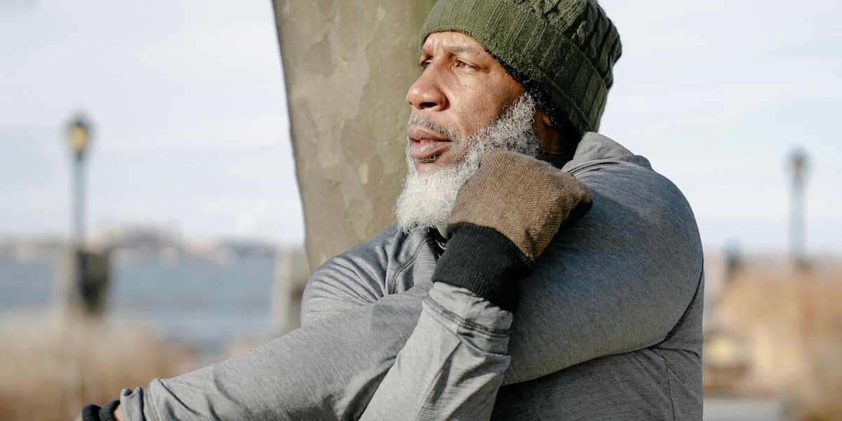 An older Black gentleman wearing a knit cap, long sleeve shirt and gloves looks away from the camera and stretches as he warms up to work out in a New York City park.