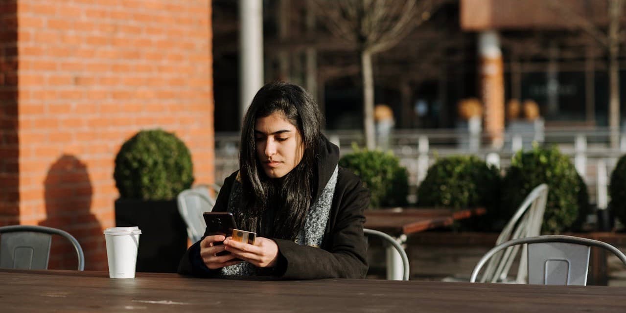 Una mujer joven de piel oscura y cabello largo y oscuro que lleva un abrigo negro de invierno se sienta afuera en la mesa de un café bajo el sol, mirando su teléfono con una tarjeta de crédito para pagar algo.
