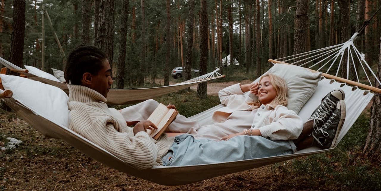 Un joven negro y una joven blanca con elegantes ropas de otoño yacen en una hamaca en medio del bosque, leyendo y sonriendo el uno al otro.