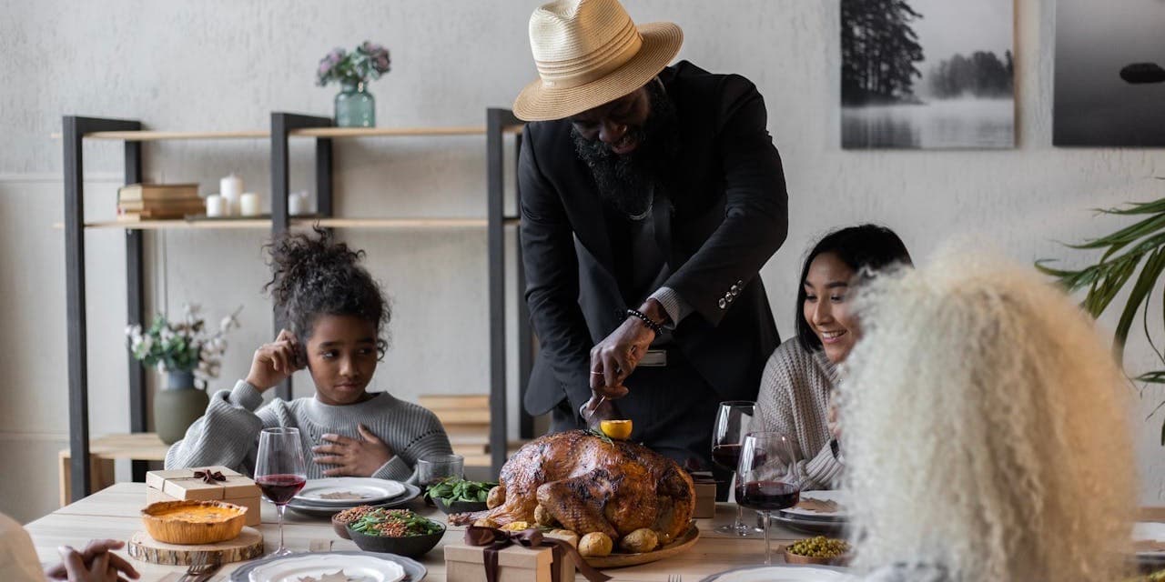 A diverse group of family and friends is gathered at a dining room table in a bright, airy room. At center, a handsome Black man wearing a black suit and hat carves a Thanksgiving turkey.