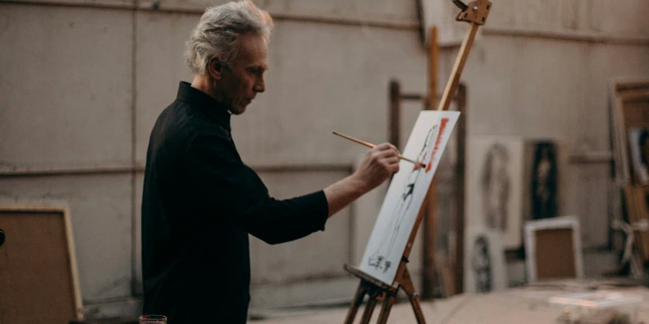 An older, thin white man with grey hair stands at an easel in a large, airy art studio, painting on a canvas.