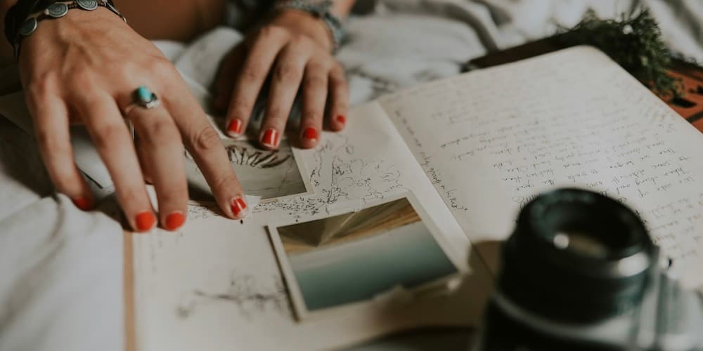 Un primer plano de las manos de una mujer blanca con joyas modernas y esmalte de uñas rojo desconchado sobre un diario con fotos y bocetos, y una cámara cerca.