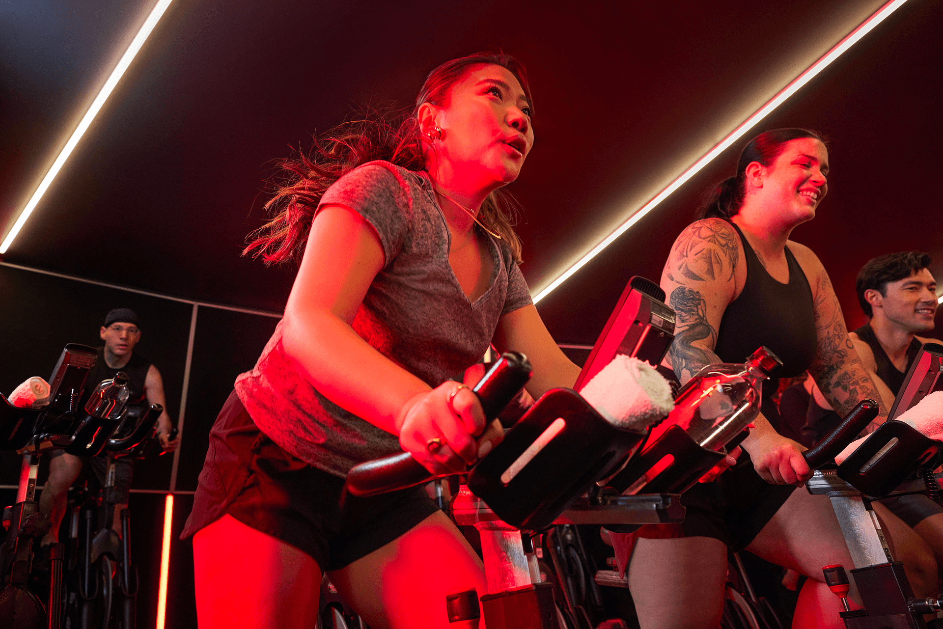 Un grupo de personas en una clase de ciclismo cubierta con poca luz, pedalean intensamente bajo una luz roja. Una mujer central, vestida con una camiseta gris, lidera la campaña, mientras que otras, incluida una mujer tatuada y sonriente, van a su lado.