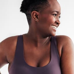 Frente a un fondo neutro, mujer vestida con ropa deportiva sujetándose la parte derecha de la cintura con ambas manos, mirando y sonriendo hacia la izquierda.