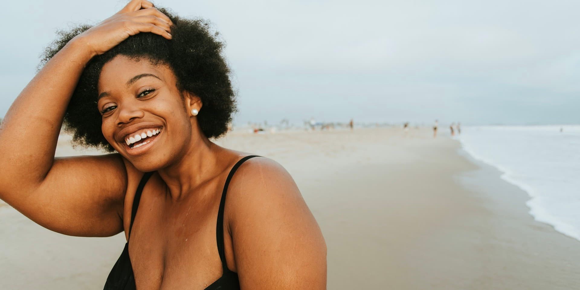Una mujer negra con curvas vestida con un traje de baño negro está de pie en la playa y sonríe de par en par a la cámara mientras se pasa los dedos por el pelo.