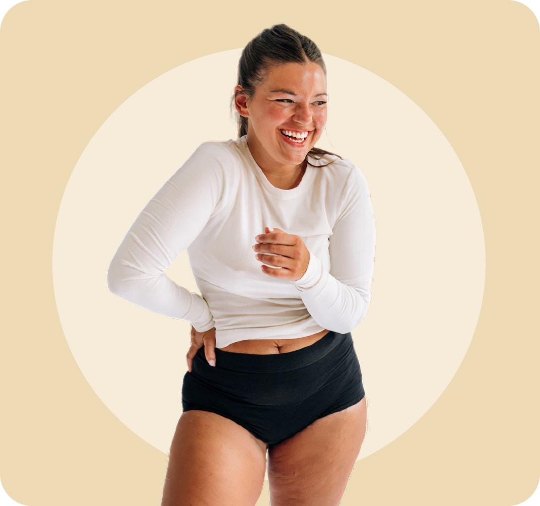 Mujer vestida con un top blanco de manga larga y ropa interior negra de cintura alta, riendo y mirando a su izquierda con una mano en la cintura y la otra sobre el estómago.