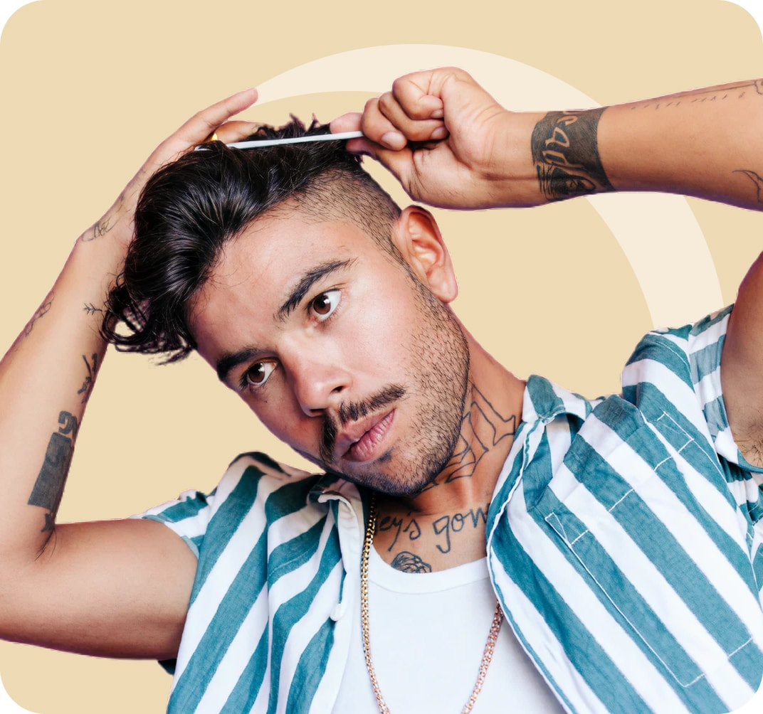 Hombre con tatuajes peinándose mientras mira ligeramente hacia abajo. Lleva una camisa a rayas sobre una camiseta blanca sin mangas y se para frente a un fondo beige.