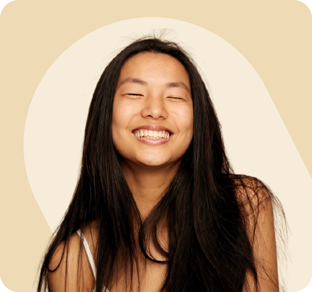 Mujer de pelo largo y negro, sonriente con los ojos cerrados frente a un fondo beige neutro. Lleva una camiseta blanca sin mangas con tirantes finos.